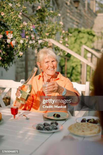 senior woman holding alcohol bottle while sitting at dining table in back yard - alcool stock pictures, royalty-free photos & images