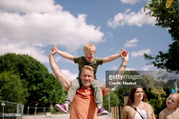 father carrying son on shoulders while walking by woman and daughter at park during sunny day - stockholm park stock pictures, royalty-free photos & images