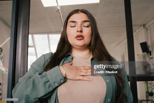 young businesswoman with eyes closed and hand on chest at office - nervosität stock-fotos und bilder