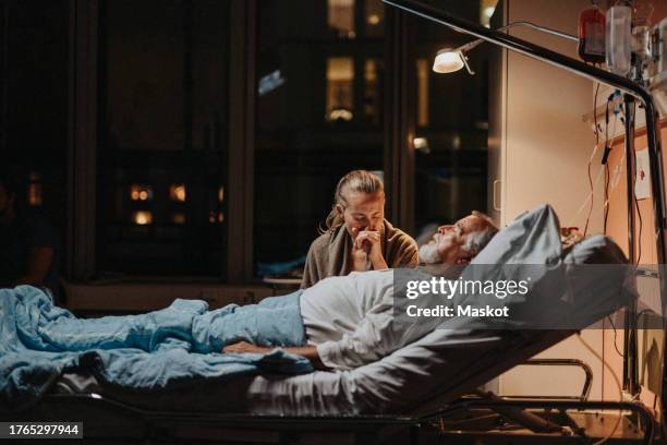 sad woman sitting near senior patient lying down on bed in intensive care unit at hospital - patient lying down stock pictures, royalty-free photos & images