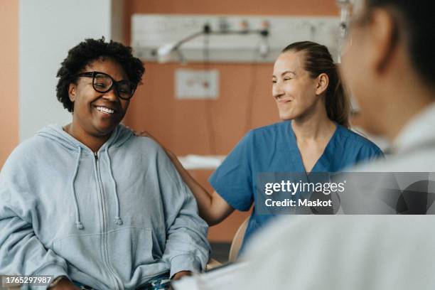 smiling female nurse consoling happy patient sitting with doctor during consultation at hospital - heilbehandlung stock-fotos und bilder