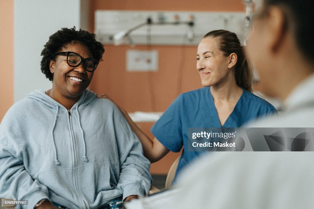 Smiling female nurse consoling happy patient sitting with doctor during consultation at hospital