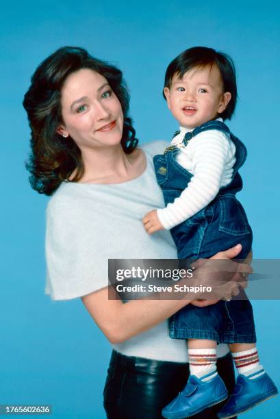 Portrait of English actress Olivia Hussey holding her son, Maximillian Fuse, as they pose against a blue background, Los Angeles, California, 1984.