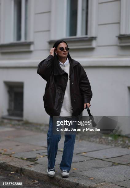 Anna Winter is seen wearing silver earrings, black rimless oval sunglasses from Balenciaga, a dark brown Letter jacket from Gant, a grey sweater with...