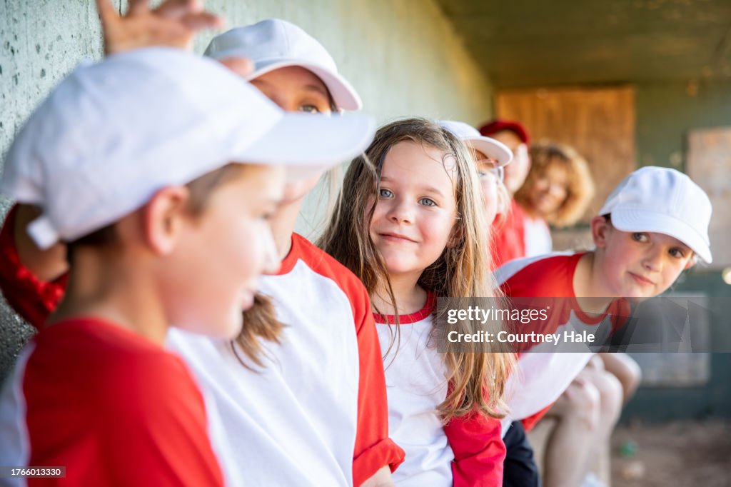 Six young children who attend elementary school are wearing baseball team uniforms as they sit in a dugout