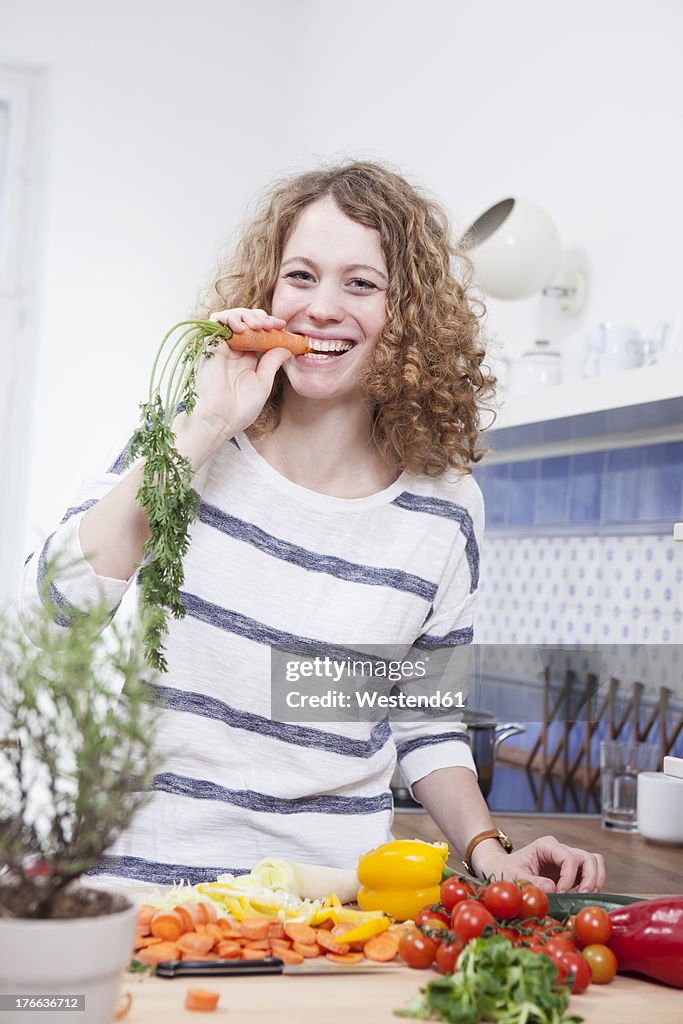 Germany, Bavaria, Munich, Young woman eating carrot in kitchen, smiling, portrait