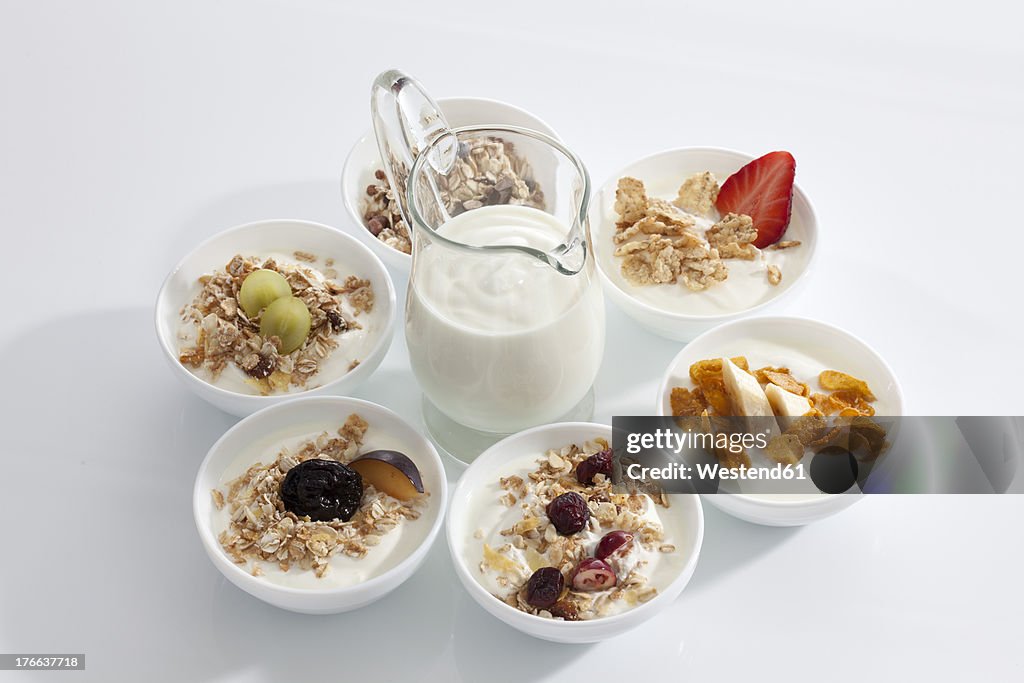 Carafe of yogurt around bowls of muesli yogurt with fruits on white background