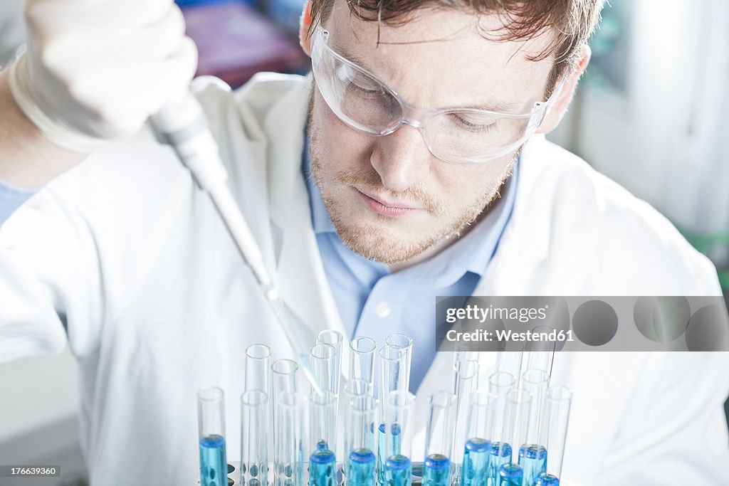 Germany, Young scientist pipetting blue liquid into test tubes, close up