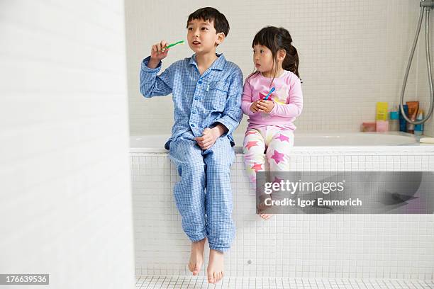 brother and sister sitting on edge of bath with toothbrushes - brothers bathroom stock-fotos und bilder