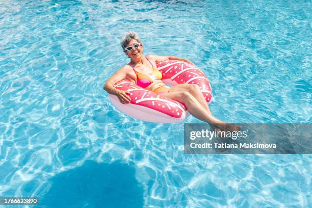 a retired woman paddles in a pink inflatable circle donut in a pool of blue water, high angle view. - schwimmring stock-fotos und bilder