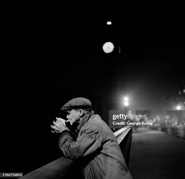 Homeless man Henry Almond leans on the parapet of Westminster Bridge and relights his cigarette, London, November 1950. The illuminated clock of Big...