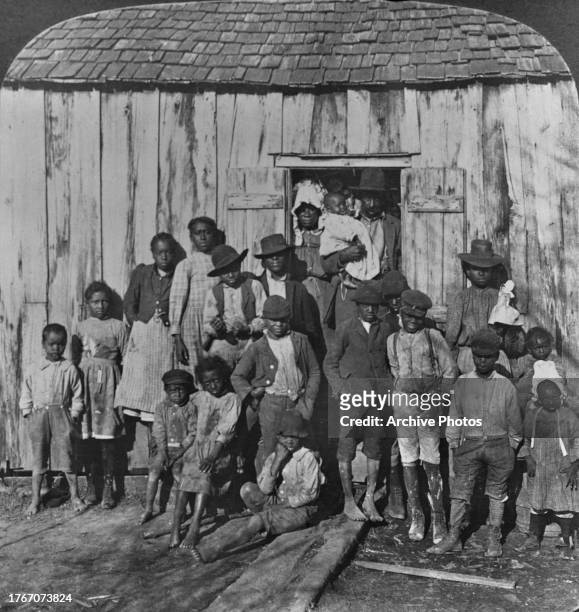 Couple stands in the doorway of their wood-built home with the father holding a child and seventeen children standing outside the family home in...