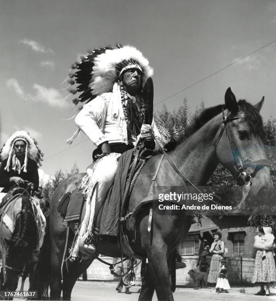 View of an unidentified man, in ceremonial attire and a feathered war bonnet, as he rides a horse during the Gallup Inter-Tribal Indian Ceremonial...
