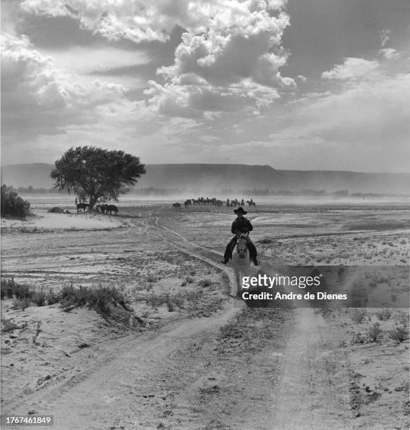 View of a man rides a horse along a dirt path, Chinle, northern Arizona, mid twentieth century. A group of men & horses are visible in the distance.