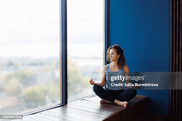 peaceful female yoga athlete finds serenity in lotus position sitting gracefully on wooden window sill - mudra stock pictures, royalty-free photos & images