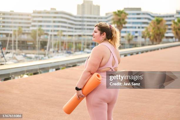 joven mujer de talla grande de pie con esterilla de yoga - voluptuoso fotografías e imágenes de stock
