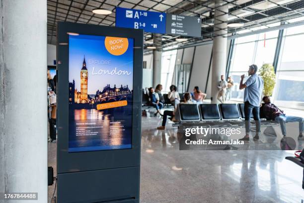 airport terminal gate with seats and a london billboard with blurred people in the background - airport display stock pictures, royalty-free photos & images