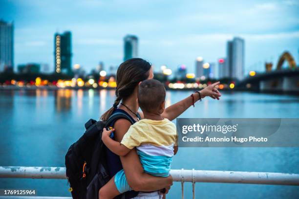 young woman with her son in her arms showing him da nang city at dusk - river han stock pictures, royalty-free photos & images