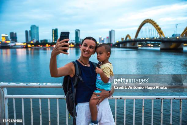 young woman with her child in her arms taking a selfie with the city of da nang in the background and the dragon bridge (cau rong) at sunset. - river han stock pictures, royalty-free photos & images