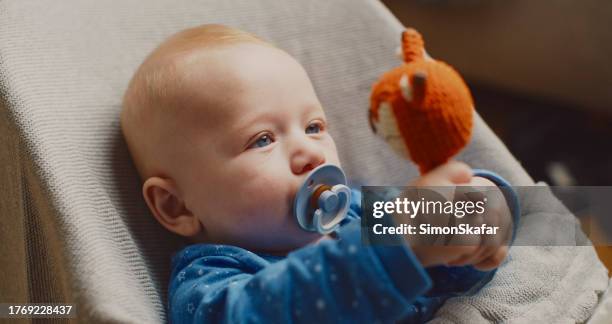 close-up of cute baby boy looking at toy while holding pacifier in mouth on bouncer at home - overall stockfoto's en -beelden