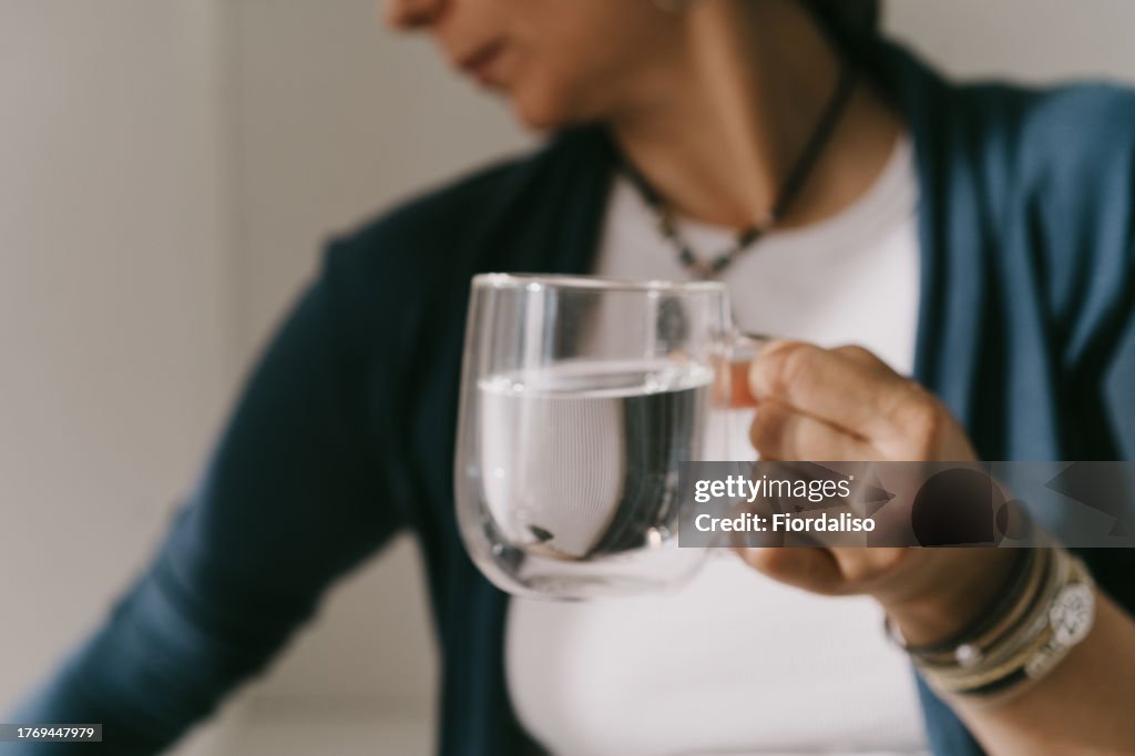 A woman standing at the kitchen counter with a clear mug or glass of water. The concept of body water balance, dehydration problems, healthy eating, taking nutritional supplements and vitamins, quenching thirst