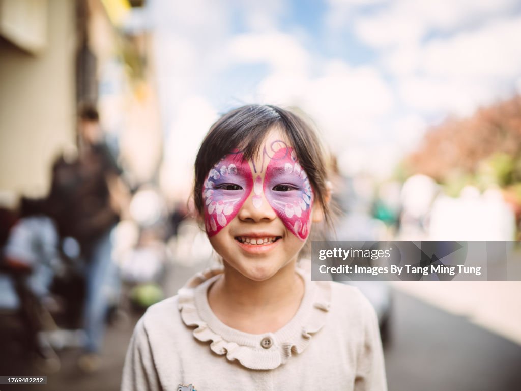 Lovely cheerful girl with beautiful face painting smiling joyfully at the camera while exploring in a theme park