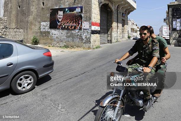 Two young Syrian soldiers ride a motorcycle past a huge poster bearing a portrait of President Bashar al-Assad next to a painting of a Syria's...
