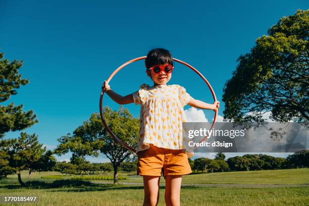 cheerful little asian girl with red sunglasses playing with a hula hoop in park on a sunny day against beautiful blue sky. outdoor fun. active and healthy lifestyle - hooping stock pictures, royalty-free photos & images