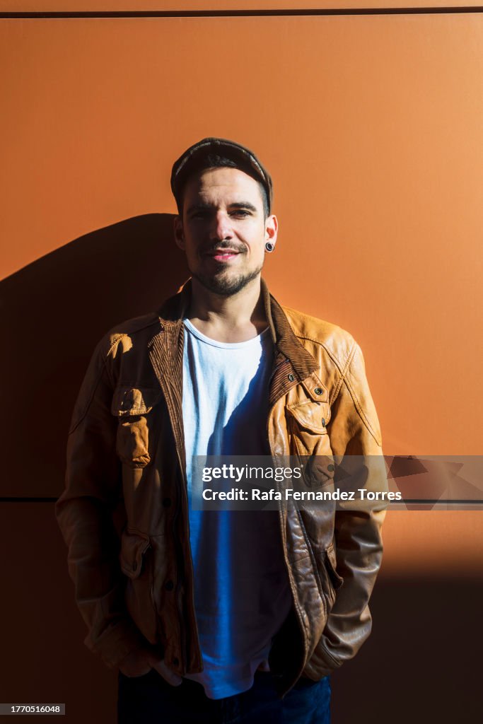 A Young handsome man in leather jacket leaning on a wall outdoor looking at camera