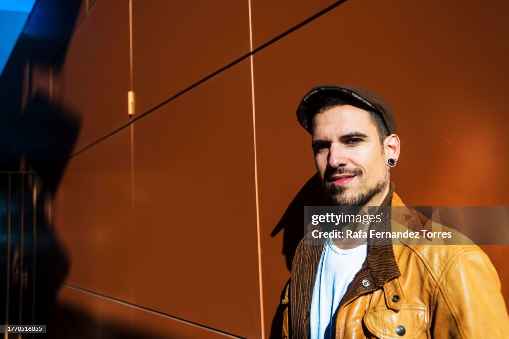 A Young handsome man in leather jacket leaning on a wall outdoor looking at camera