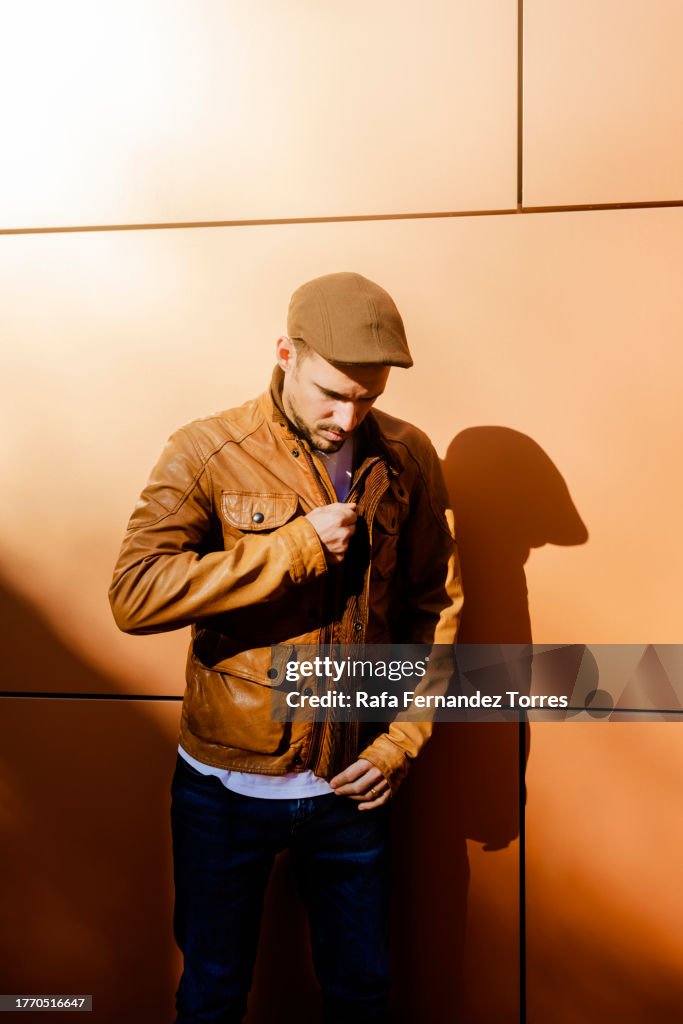 Attractive young man in leather jacket and cap, walking by business building, buttoning his jacket.