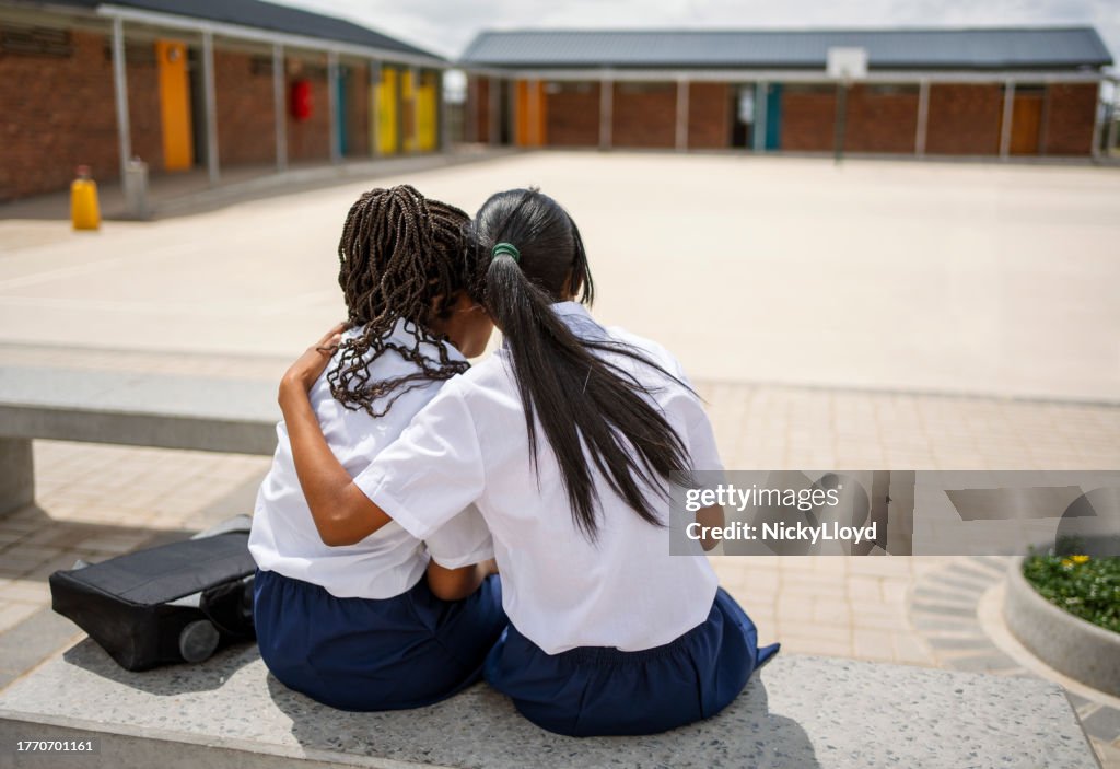 Two schoolgirls sitting together on bench in school campus during break