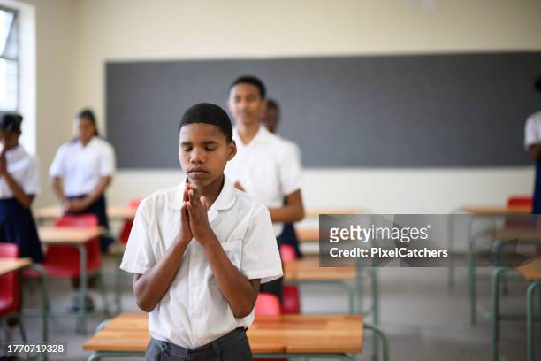 front view schoolboy standing praying in classroom with schoolmates - child praying school stock pictures, royalty-free photos & images