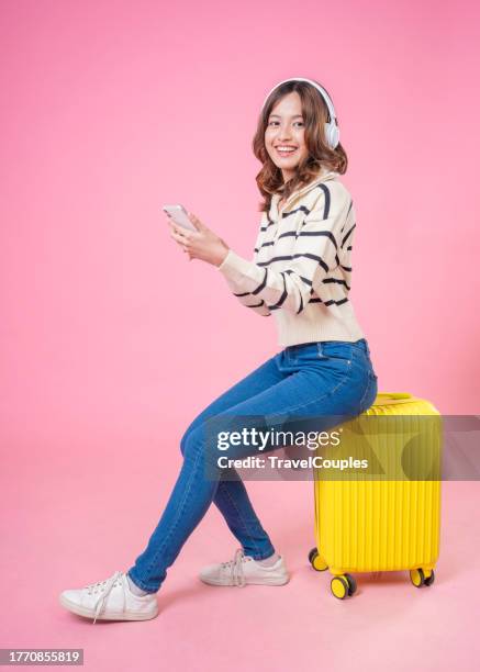 happy asian woman traveler listening to the music in the headphones with luggage isolated on pink background. tourist girl having cheerful holiday trip concept. - push cart stock pictures, royalty-free photos & images