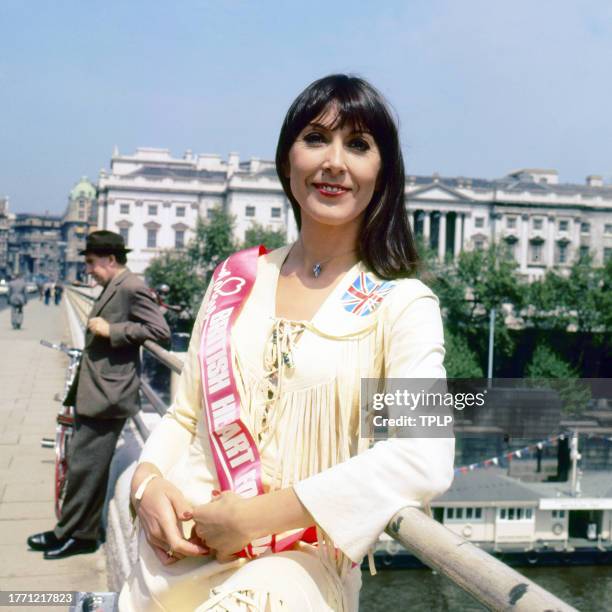 Portrait of British singer and actress Anita Harris, with a sash that reads 'British Heart Foundation,' as she stands on Waterloo Bridge, London,...