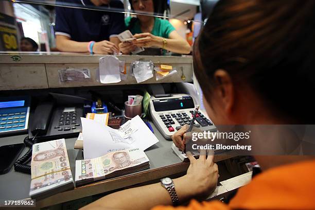 An employee serves customers as bundles of Thai one-thousand baht banknotes sit on a bench at a Super Rich 1965 Co. Currency exchange store in...