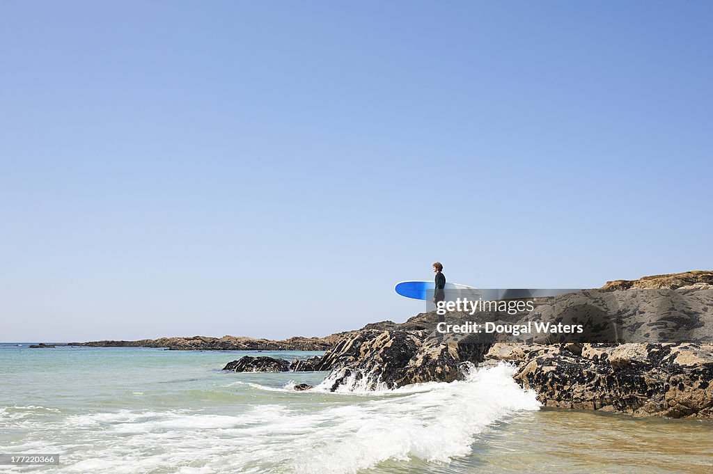 Surfer standing on rocks and looking out to sea.