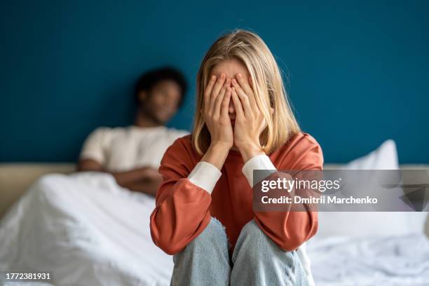 crying woman closing face by hands sitting in bed, frustrated man lying on background. - human relationship stock pictures, royalty-free photos & images