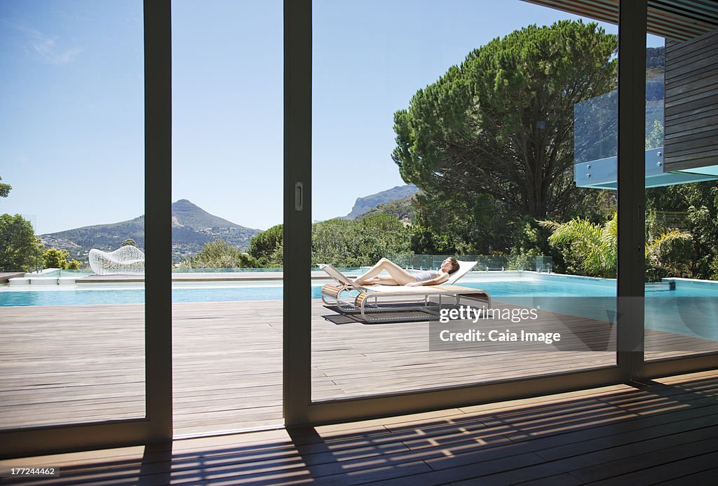 Woman sunbathing on lounge chair next to luxury swimming pool with mountain view