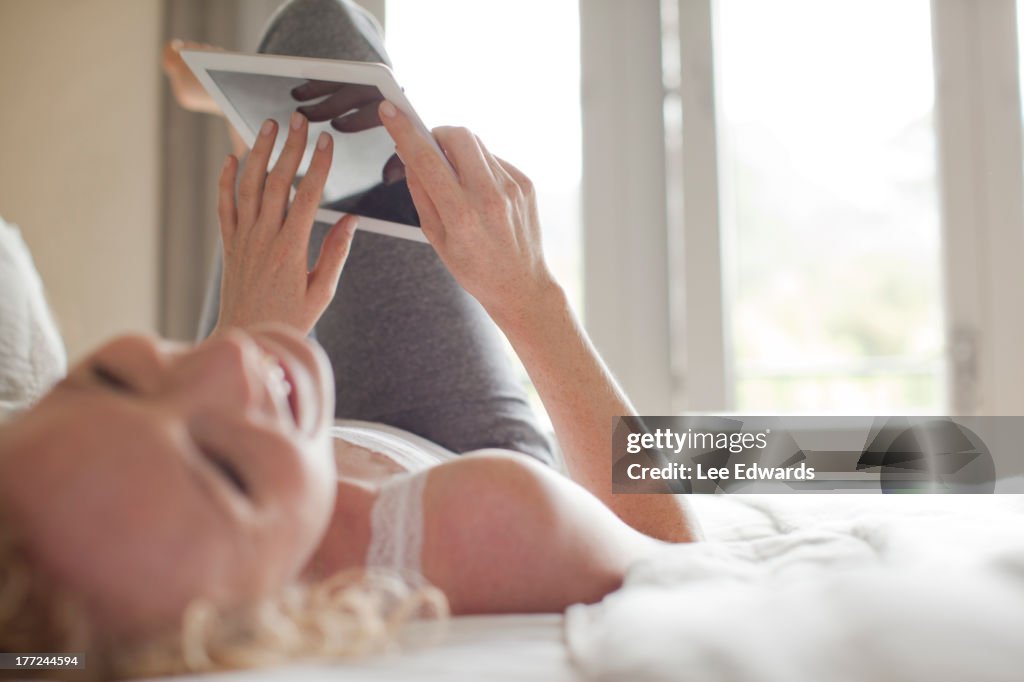 Laughing woman laying in bed using digital tablet