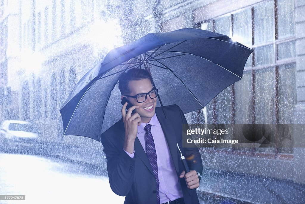 Happy businessman talking on cell phone under umbrella in rainy street