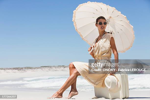 beautiful woman sitting on a suitcase with an umbrella on the beach - a woman wear hat and sunglasses stock pictures, royalty-free photos & images