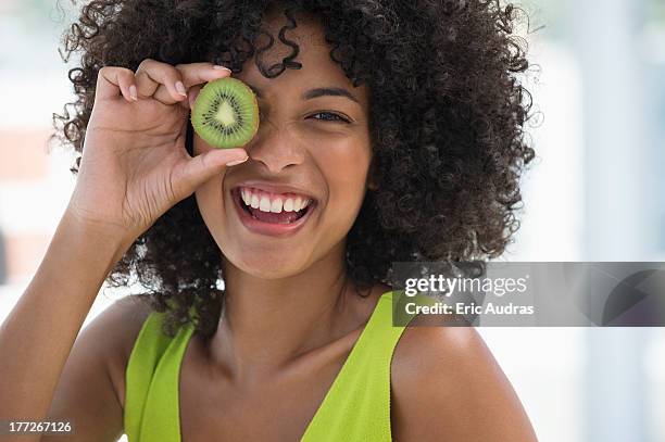 smiling woman holding a kiwi fruit in front of her eye - kiwi fruit stock pictures, royalty-free photos & images