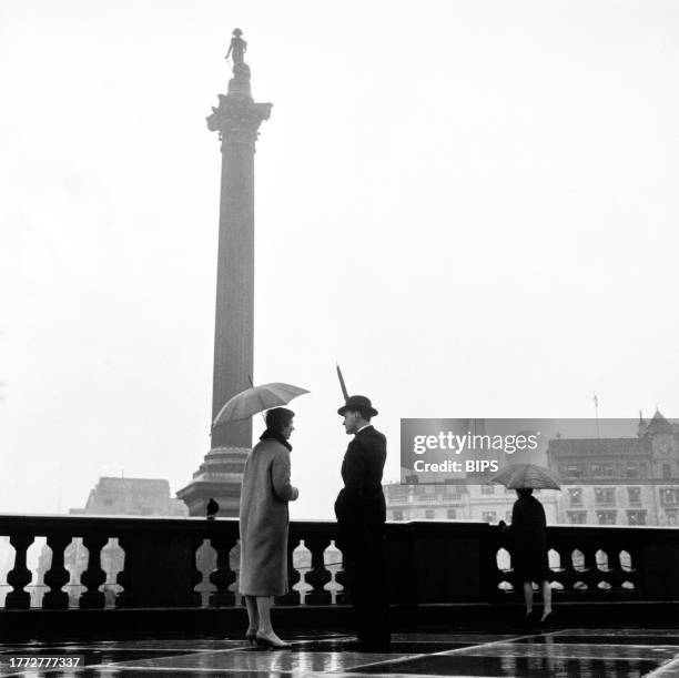 Woman sheltering beneath an umbrella as a man in a bowler hat points his rolled-up umbrella at Nelson's Column, a granite Corinthian column...