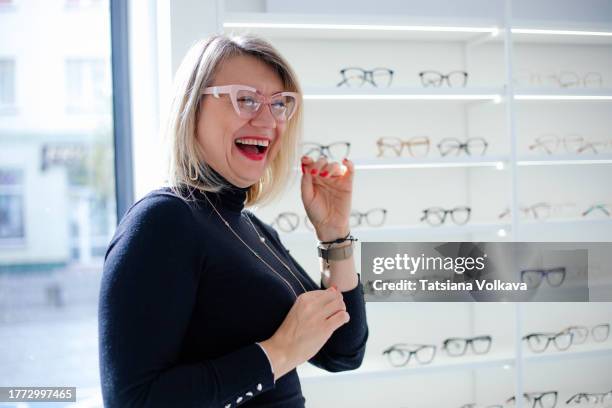 happy blonde female client trying on pair of glasses in brightly illuminated optician store - gafas de ver fotografías e imágenes de stock