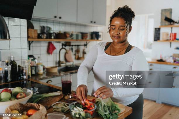 smiling young woman preparing salad bowl in kitchen at home - comida sana fotografías e imágenes de stock