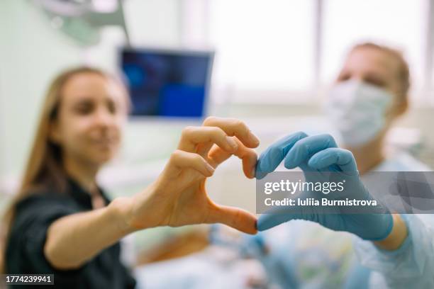 experienced female dentist and charming young patient sitting in dentist office making heart shape with hands - guante quirúrgico fotografías e imágenes de stock