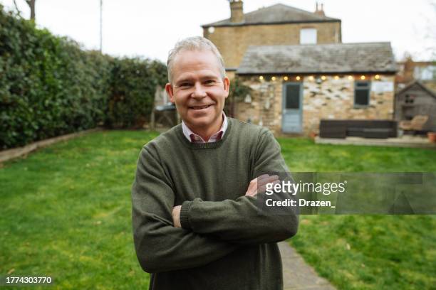 senior homeowner in uk with arms crossed standing in backyard, looking at camera and smiling - residential building photos stock pictures, royalty-free photos & images