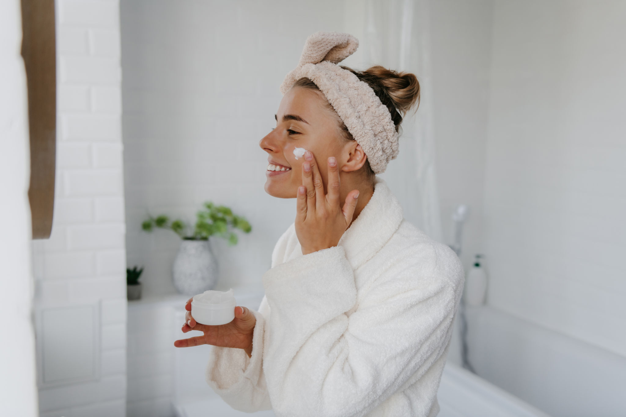 Happy young woman in bathrobe applying face cream while standing in bathroom Happy young woman in bathrobe applying face cream while standing in bathroom