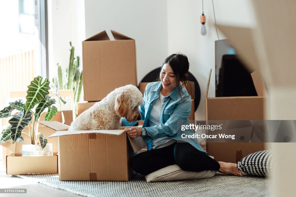 Young Asian woman having fun with her dog while unpacking cardboard boxes in the living room
